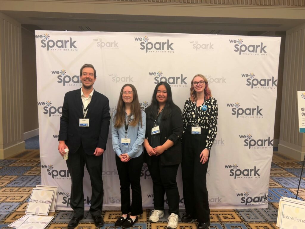 A photo of four people standing in front of a backdrop with Sarah Wilkins to the far right
